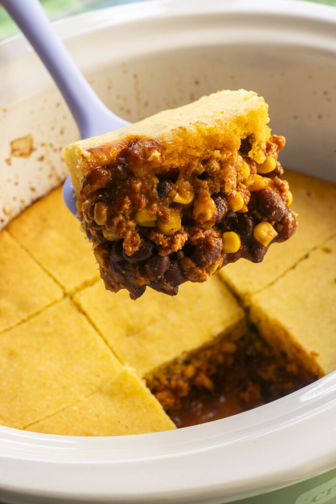 A purple spatula lifting a square of enchilada casserole, revealing the saucy ground turkey, corn, and black bean filling underneath the cornbread.