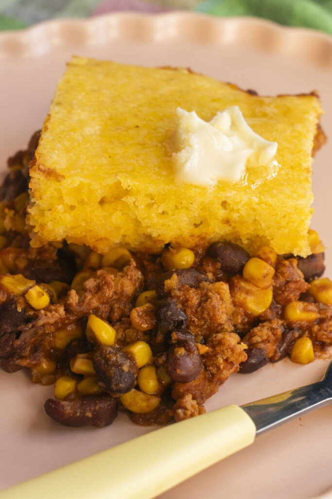 A square of cornbread-topped enchilada casserole served on a pink plate with a pat of melting butter.