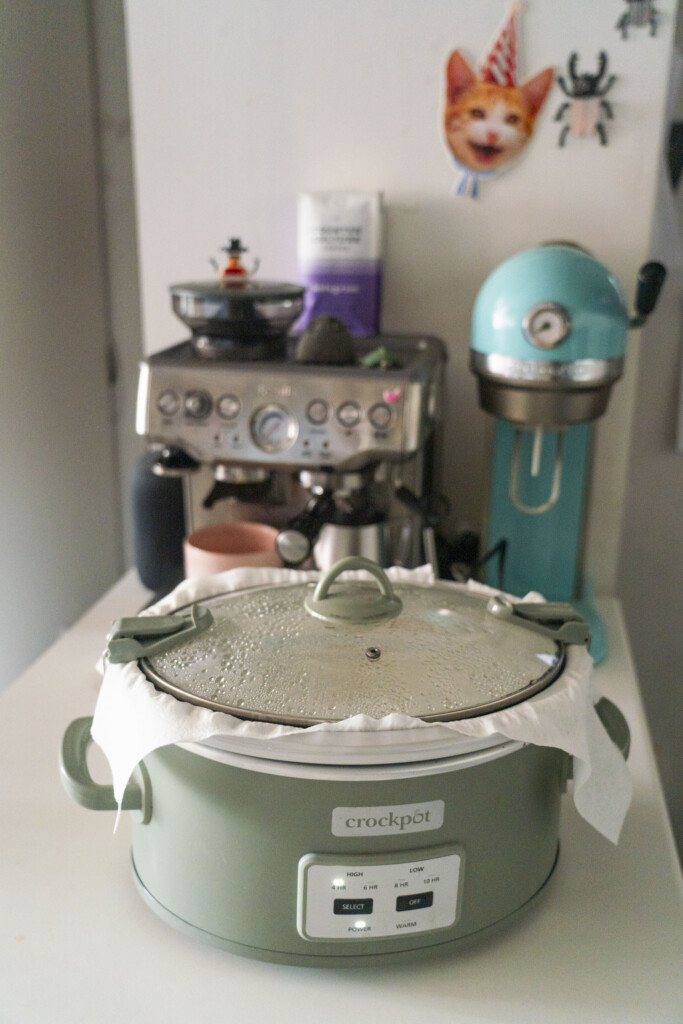A green slow cooker covered with a clean white kitchen towel under the lid to absorb moisture while the cornbread bakes.