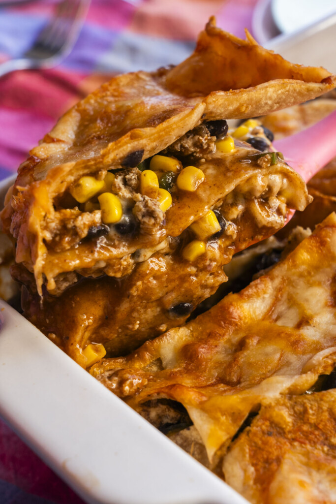 A close-up of a cheesy enchilada being lifted from a baking dish, showing melted cheese, ground meat, corn, and black beans inside, with a colorful cloth in the background.