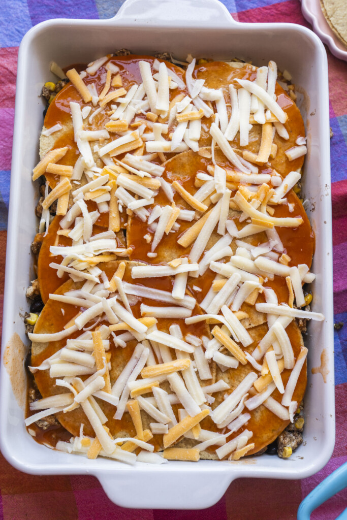 A white baking dish filled with layers of tortillas, sauce, and shredded cheese, sitting on a colorful tablecloth. The dish is ready to be baked.