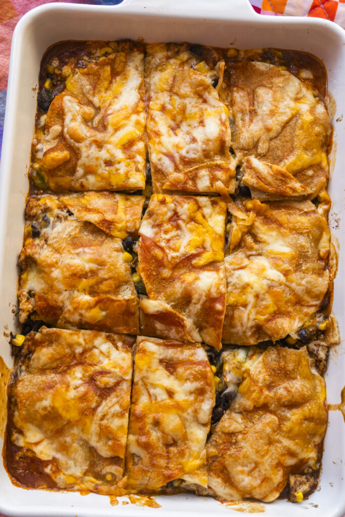 A rectangular baking dish filled with a cheesy, baked casserole cut into nine pieces. The top layer is golden brown with melted cheese, and some of the filling like beans and vegetables is visible at the edges.