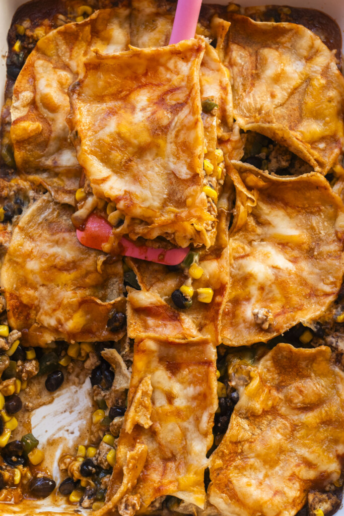 A casserole dish filled with cheesy enchilada layers, black beans, corn, and tortillas. A pink spatula lifts a serving, revealing the filling underneath the melted cheese and sauce.
