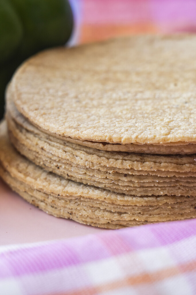 A close-up of a stack of corn tortillas on a pink and orange plaid cloth, with a blurred green pepper in the background.