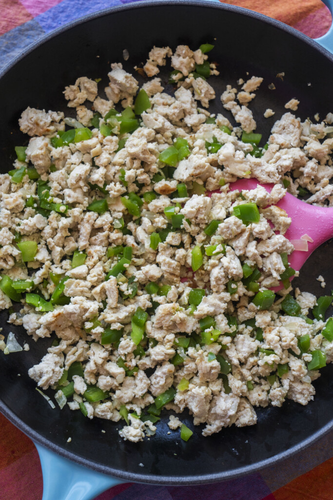 Ground turkey cooked with chopped green bell peppers and onions in a black skillet, being stirred with a pink spatula.