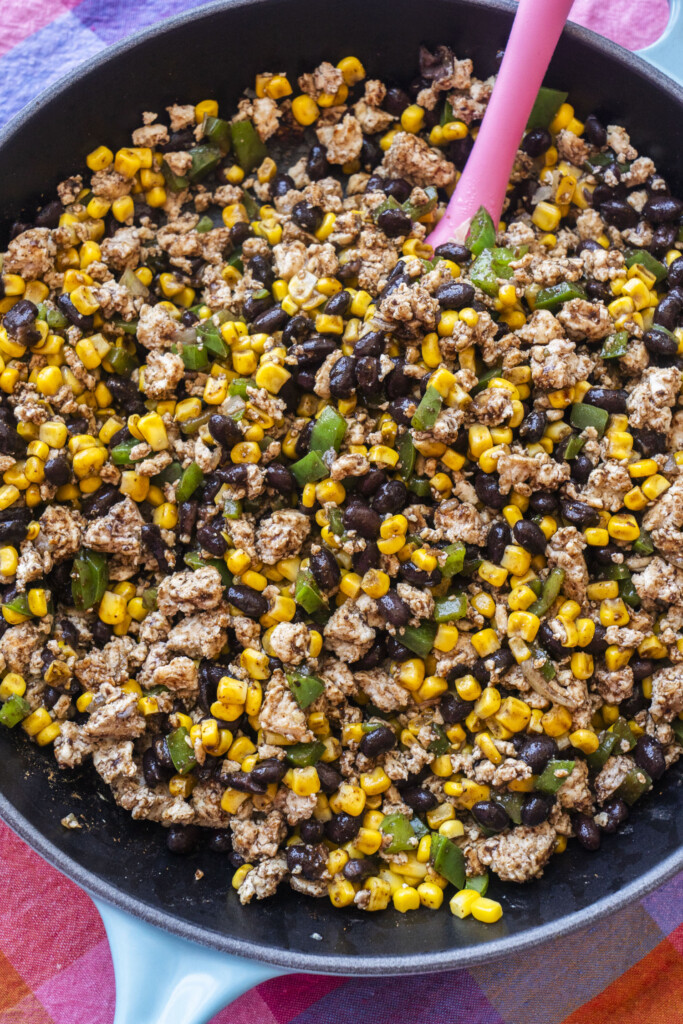 A skillet filled with a colorful mixture of ground meat, black beans, corn, and diced green bell peppers, being stirred with a pink spoon. The pan rests on a multicolored cloth.