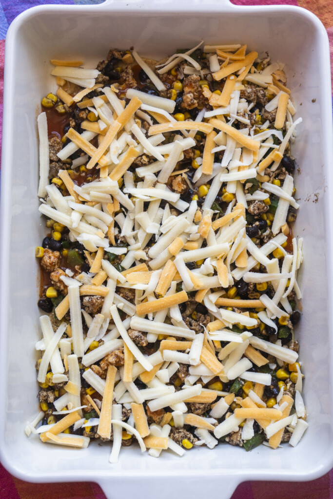 A rectangular baking dish filled with layers of shredded cheese, black beans, corn, ground meat, and vegetables, ready to be baked.