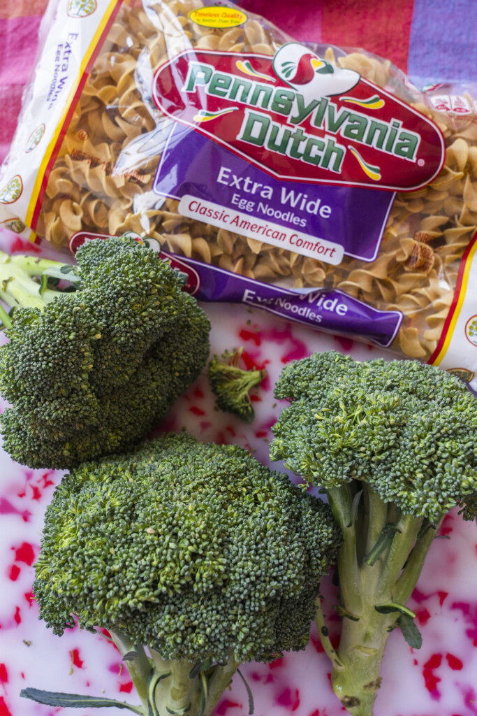 A package of Pennsylvania Dutch extra wide egg noodles sits on a surface next to three fresh broccoli heads on a white cutting board with red speckles.