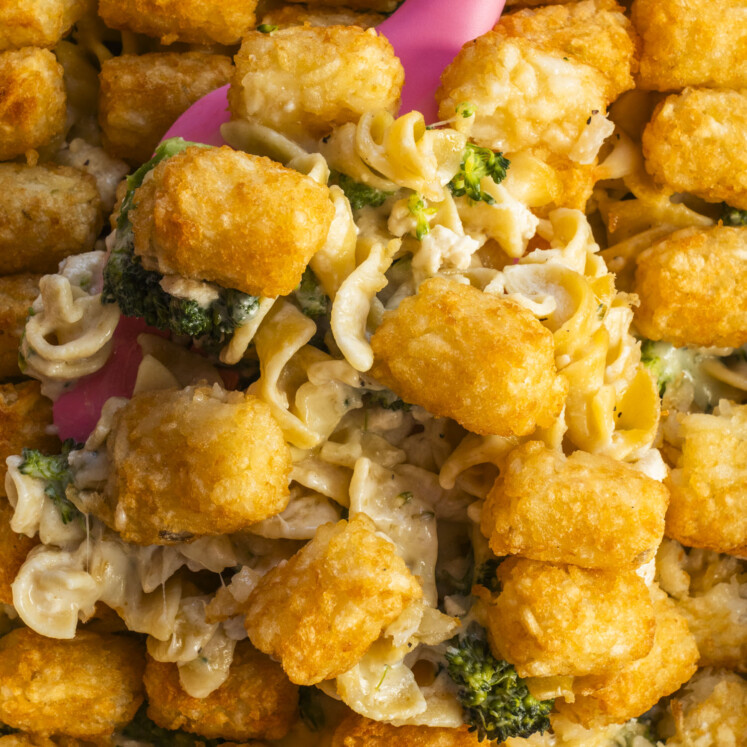 A close-up of a casserole with golden tater tots, creamy noodles, broccoli, and sauce, being served with a pink spatula.