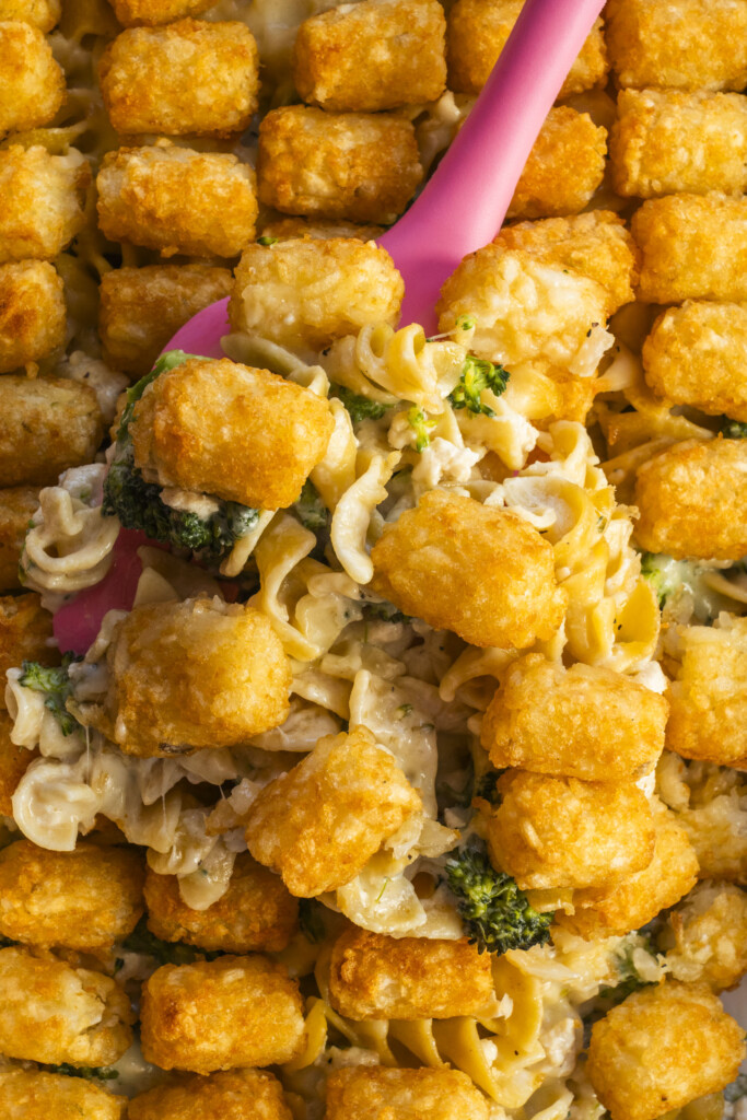 A close-up of a casserole topped with rows of golden tater tots, with a pink spoon lifting a serving to reveal creamy pasta and broccoli underneath.