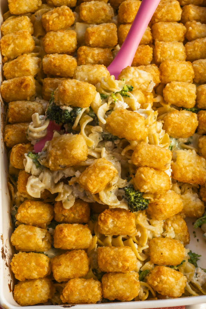 A casserole dish filled with golden tater tots on top of a creamy noodle and broccoli mixture, with a pink spatula lifting a portion.