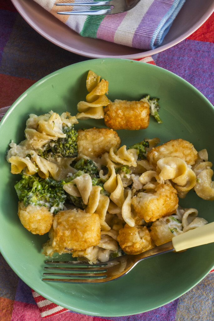 A green bowl with a serving of broccoli, egg noodles, tater tots, and creamy sauce, with a fork resting on the plate. A colorful striped napkin and a bowl with cutlery are in the background.