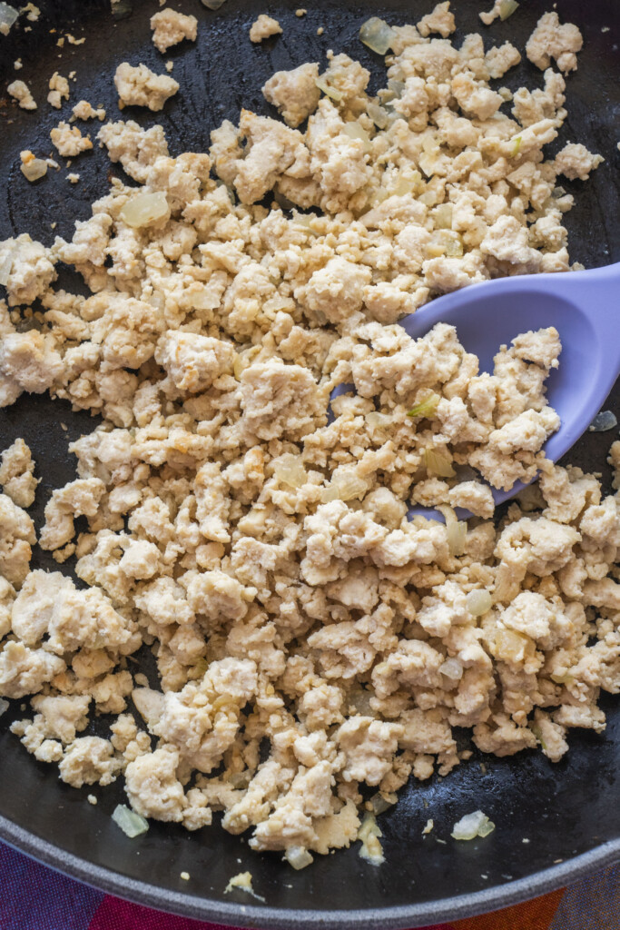 Crumbled ground chicken being cooked in a skillet with small pieces of onion, stirred with a purple spatula.