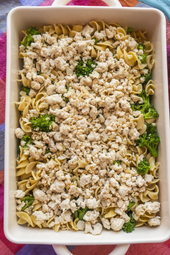 A baking dish filled with egg noodles, broccoli florets, and seasoned crumbled white meat, arranged in layers and ready to be cooked.