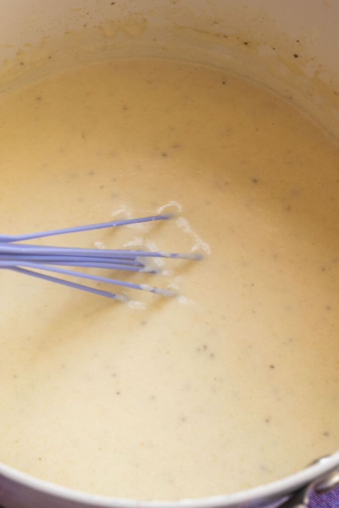 A blue whisk stirring a creamy, light orange cheese sauce in a pot.