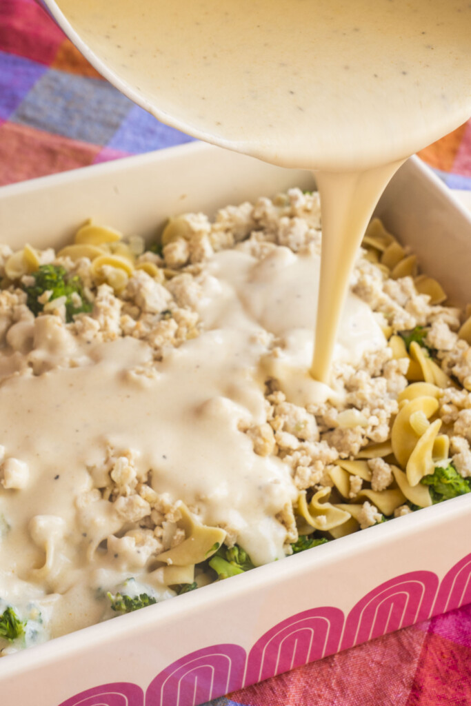 Creamy sauce being poured over a casserole dish filled with noodles, chopped greens, and crumbled meat, ready to be baked. The dish sits on a colorful, checkered tablecloth.