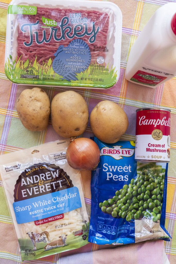 A flat lay of ingredients for a Midwest ground turkey potato casserole resting on a colorful plaid cloth, including a package of ground turkey, three whole potatoes, a yellow onion, shredded sharp white cheddar cheese, frozen sweet peas, milk, and a can of cream of mushroom soup.