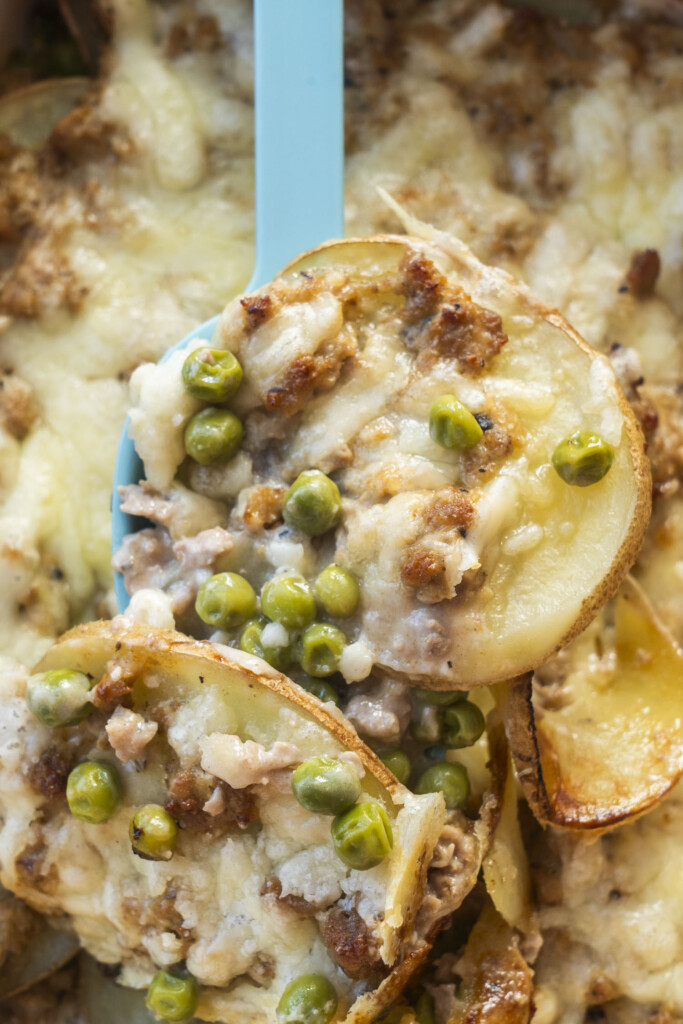 A close-up of a blue serving spoon holding a generous scoop of cheesy potato and ground turkey casserole, highlighting the melted cheese, bright green peas, and roasted potato edges.