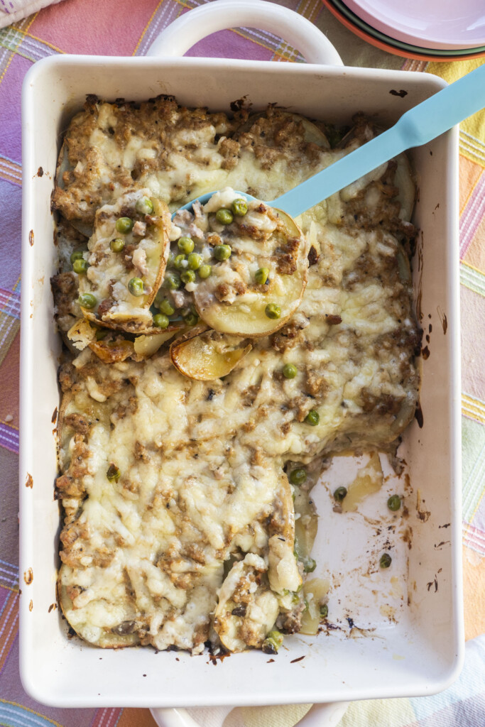 A top-down view of a freshly baked Midwest Ground Turkey Potato Casserole in a white baking dish, with a blue serving spoon lifting a hearty scoop to reveal layers of potatoes, meat, peas, and cheese.
