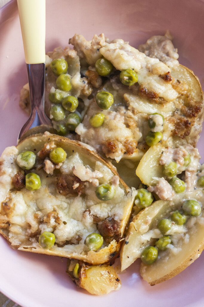 A close-up of a serving of Midwest Ground Turkey Potato Casserole in a pink bowl, showing layered sliced potatoes, savory ground turkey, green peas, and melted cheese, with a fork resting on the edge.