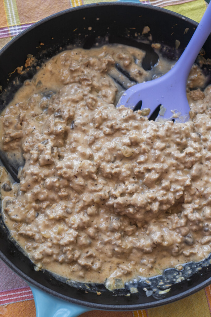A rich, creamy mixture of browned ground turkey and cream of mushroom soup simmering in a skillet with a purple spatula, ready to be poured over sliced potatoes.