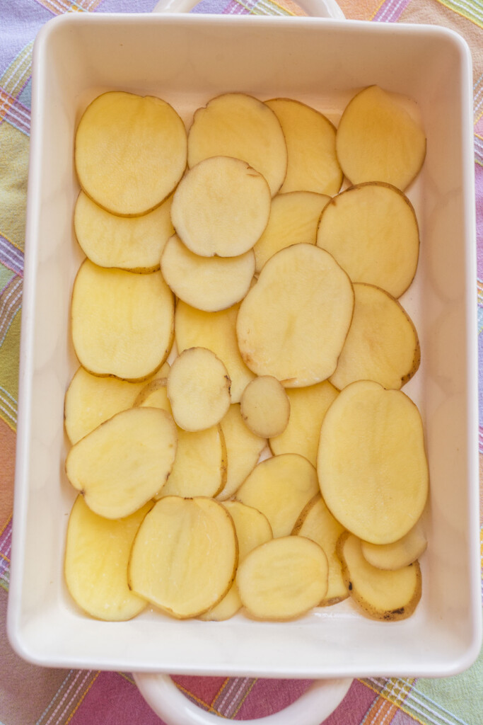 A top-down view of a white rectangular baking dish lined with an even layer of thinly sliced raw yellow potatoes, the base for a comforting Midwest ground turkey casserole.
