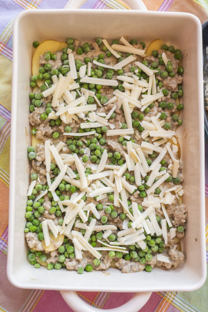 A process shot of assembling a ground turkey casserole, showing a creamy meat mixture topped with bright green peas and shredded white cheese over a layer of sliced potatoes.