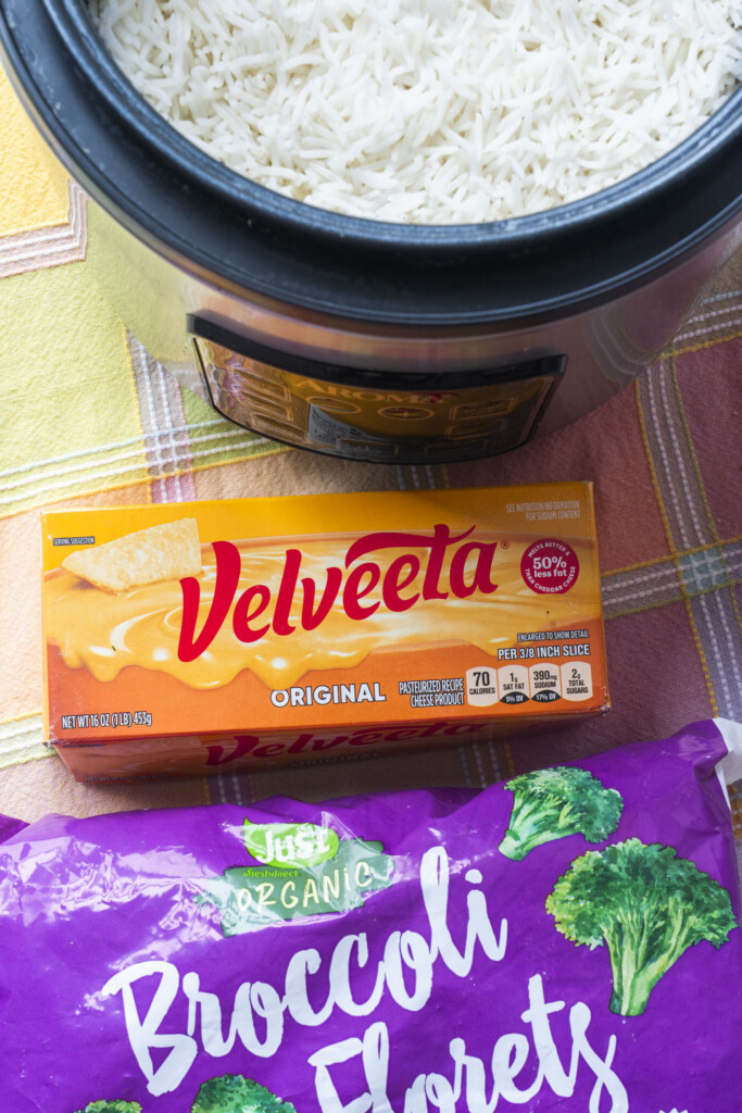 Cooked white rice in a rice cooker, a box of Velveeta cheese, and a bag of frozen broccoli florets for a casserole recipe.