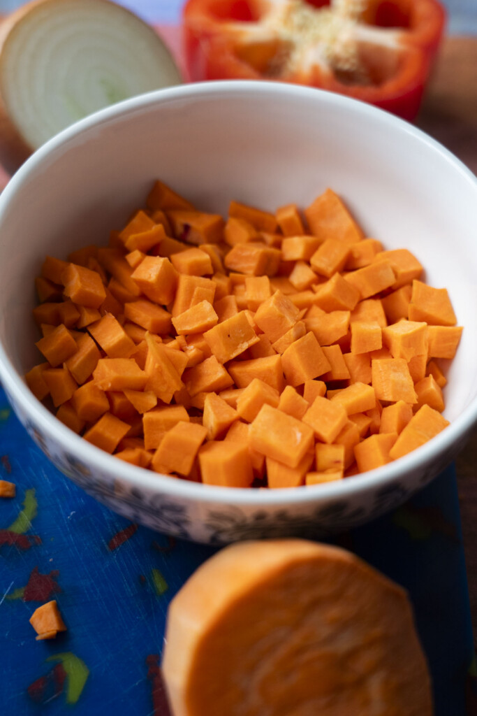 A white bowl filled with diced sweet potatoes sits on a table, surrounded by a chopped onion, a sliced red bell pepper, and a partially visible sweet potato.