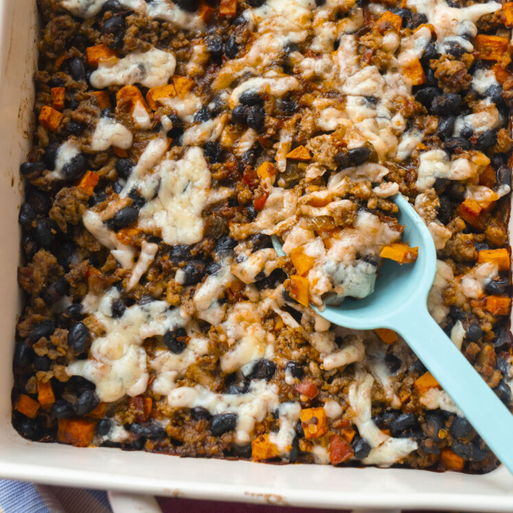 A close-up of a casserole dish filled with a baked mixture of black beans, ground meat, sweet potatoes, and melted cheese. A blue spoon is scooping out a portion from the casserole.