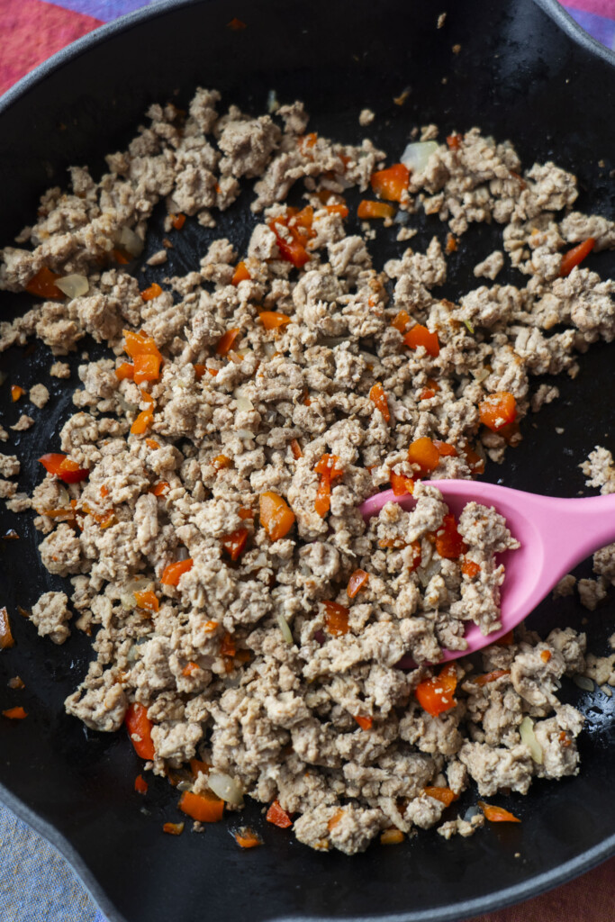 Ground meat sautéed with chopped red bell peppers and onions in a black skillet, being stirred with a pink spatula.