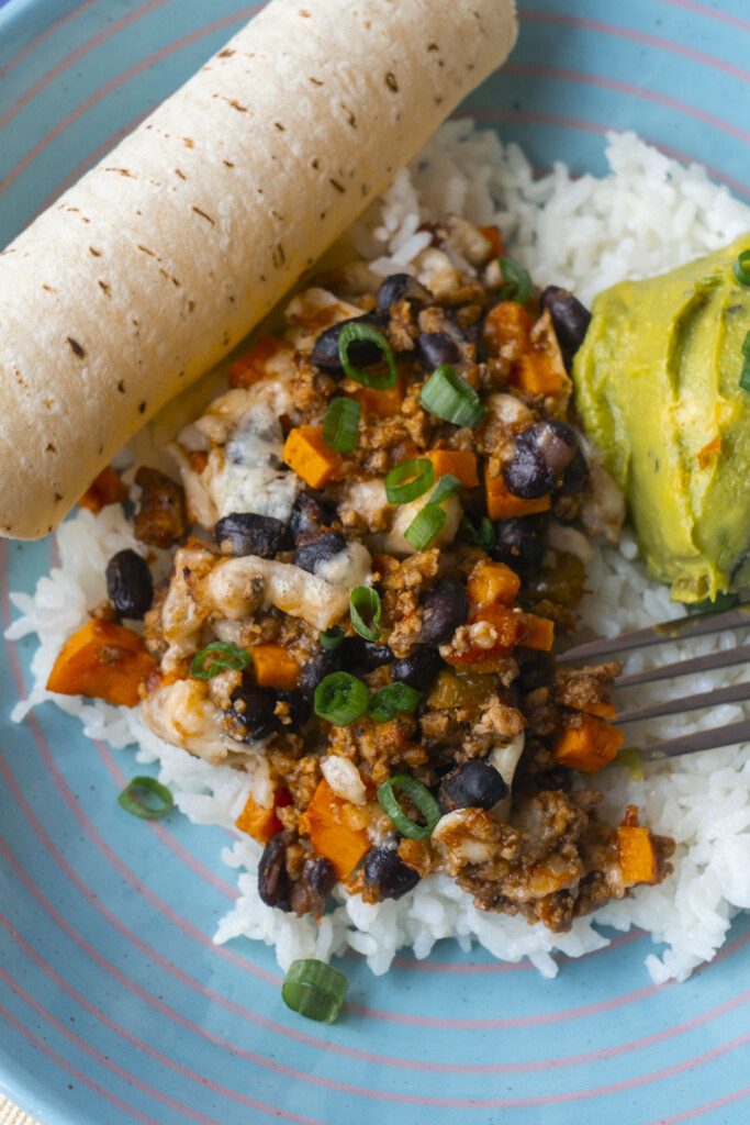 A plate with white rice topped with ground meat, black beans, diced sweet potatoes, melted cheese, and chopped green onions, served with a scoop of guacamole and a rolled tortilla.