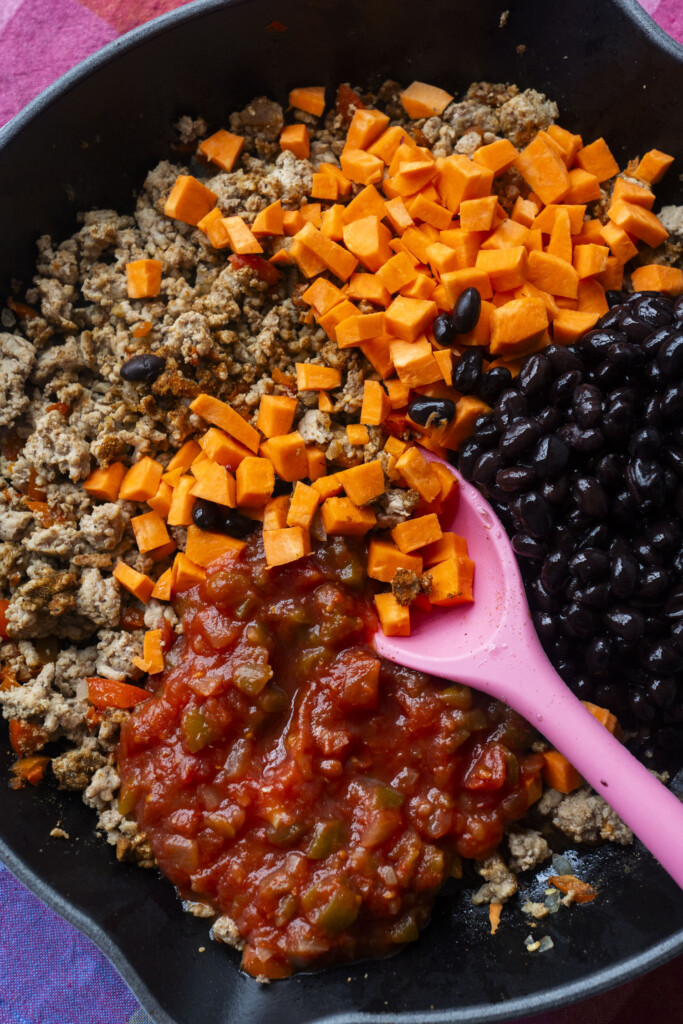 A skillet containing cooked ground meat, diced sweet potatoes, black beans, and chunky salsa, with a pink spoon resting inside.
