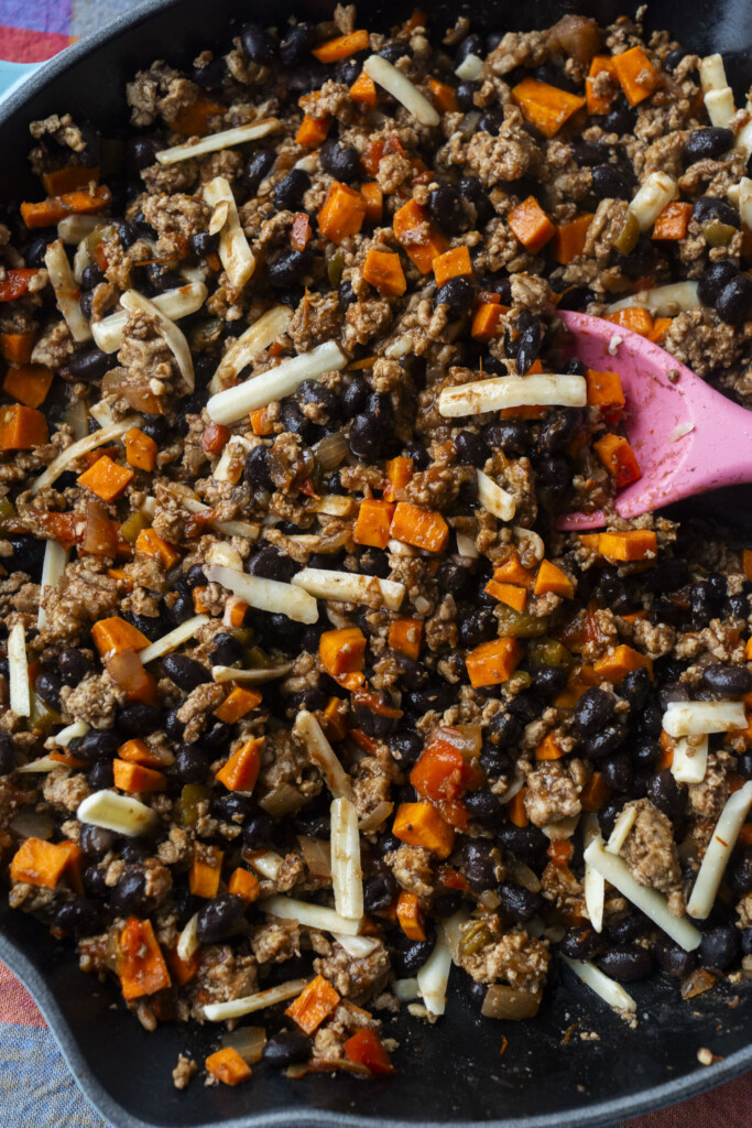 A skillet filled with a cooked mixture of ground meat, black beans, diced sweet potatoes, shredded cheese, and vegetables being stirred with a pink spatula.