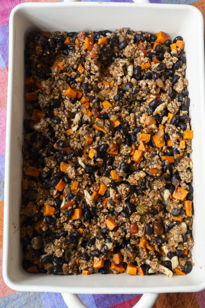 A rectangular white baking dish filled with a mixture of ground meat, black beans, diced sweet potatoes, onions, and seasonings, seen from above. The dish sits on a colorful checkered cloth.
