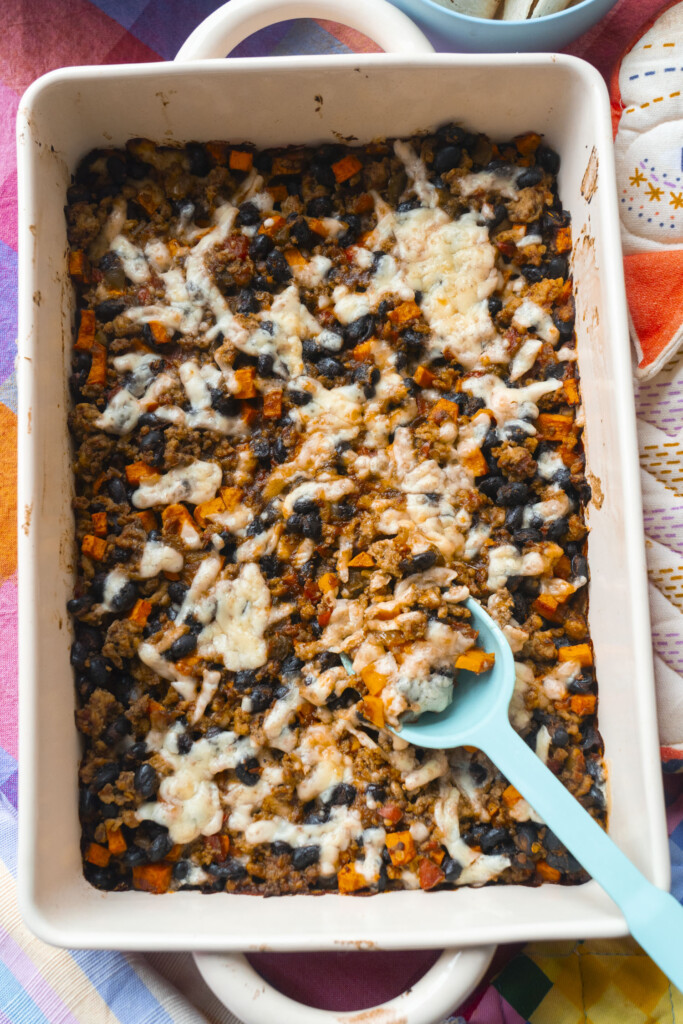 A white baking dish filled with a baked casserole featuring melted cheese, black beans, diced sweet potatoes, and grains. A turquoise serving spoon rests inside the dish, ready to serve.