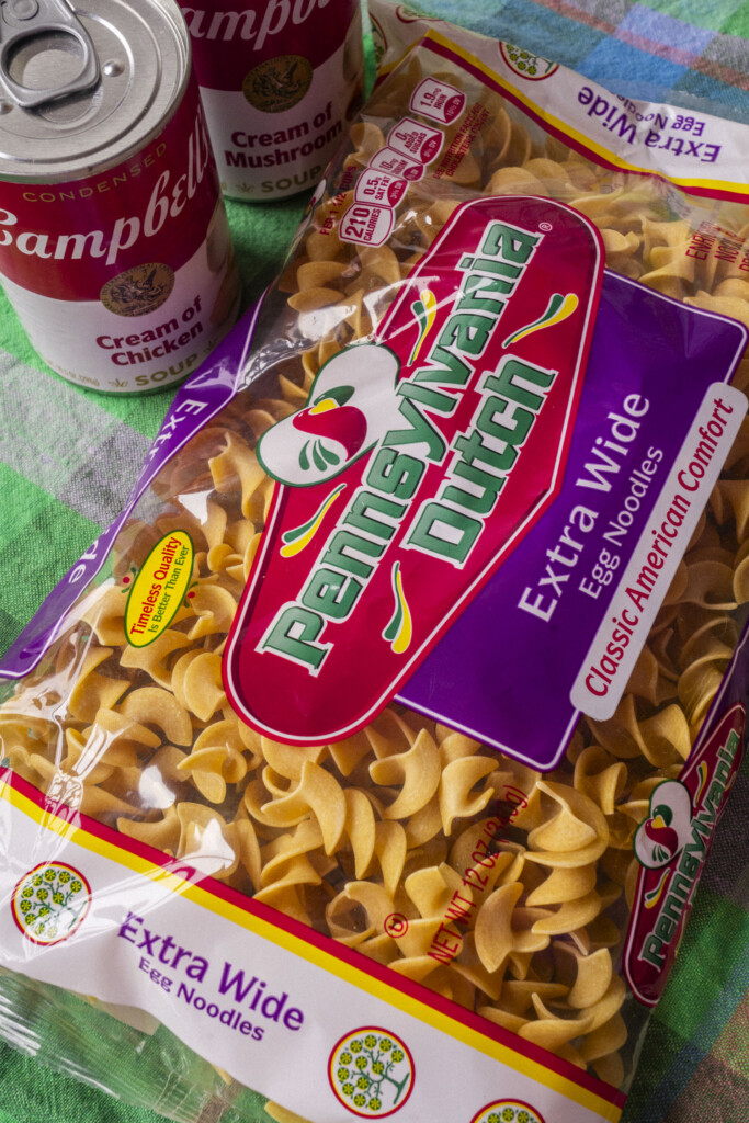 A close-up of two bags of Pennsylvania Dutch extra wide egg noodles and two cans of Campbell’s soup, one cream of chicken and one cream of mushroom, on a green and purple checkered cloth.