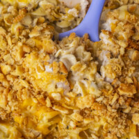 Close-up of a cheesy casserole with a golden, crunchy topping being scooped with a blue spoon. The dish contains pasta and creamy sauce, with visible melted cheese and cracker crumbs.
