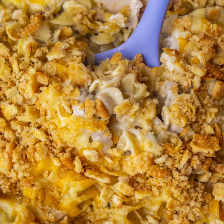 Close-up of a cheesy casserole with a golden, crunchy topping being scooped with a blue spoon. The dish contains pasta and creamy sauce, with visible melted cheese and cracker crumbs.