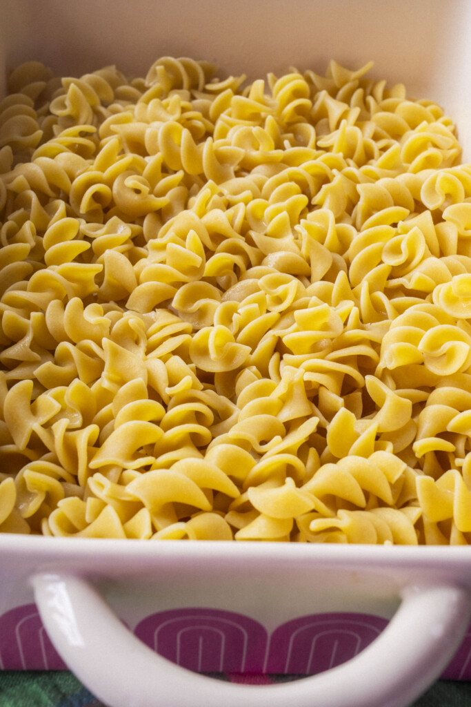 A close-up of cooked egg noodles in a white casserole dish with a pink and purple patterned side and a white handle.