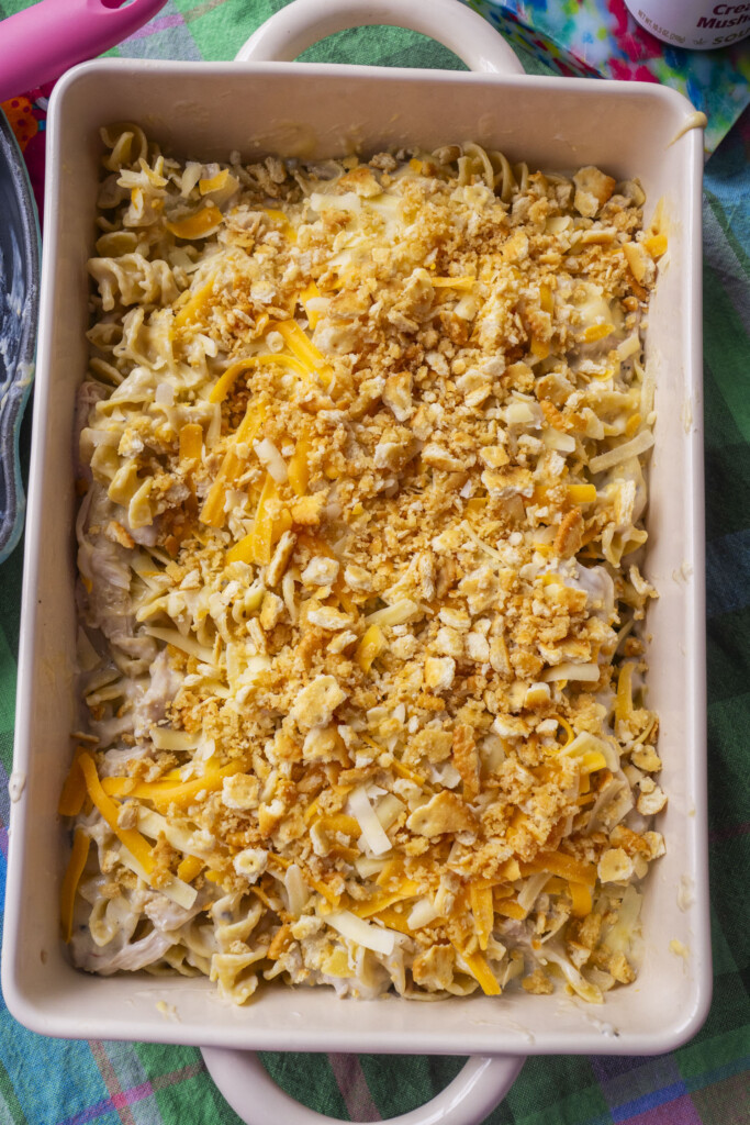A rectangular casserole dish filled with a baked noodle casserole topped with melted cheese and crushed crackers, on a colorful plaid tablecloth.