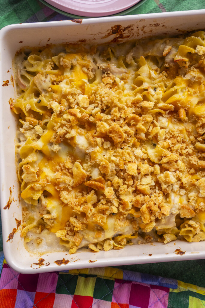 A baked Million Dollar Chicken Casserole in a white dish, topped with melted cheese and a golden, crunchy cracker crumb topping, sits on a colorful patchwork quilt.