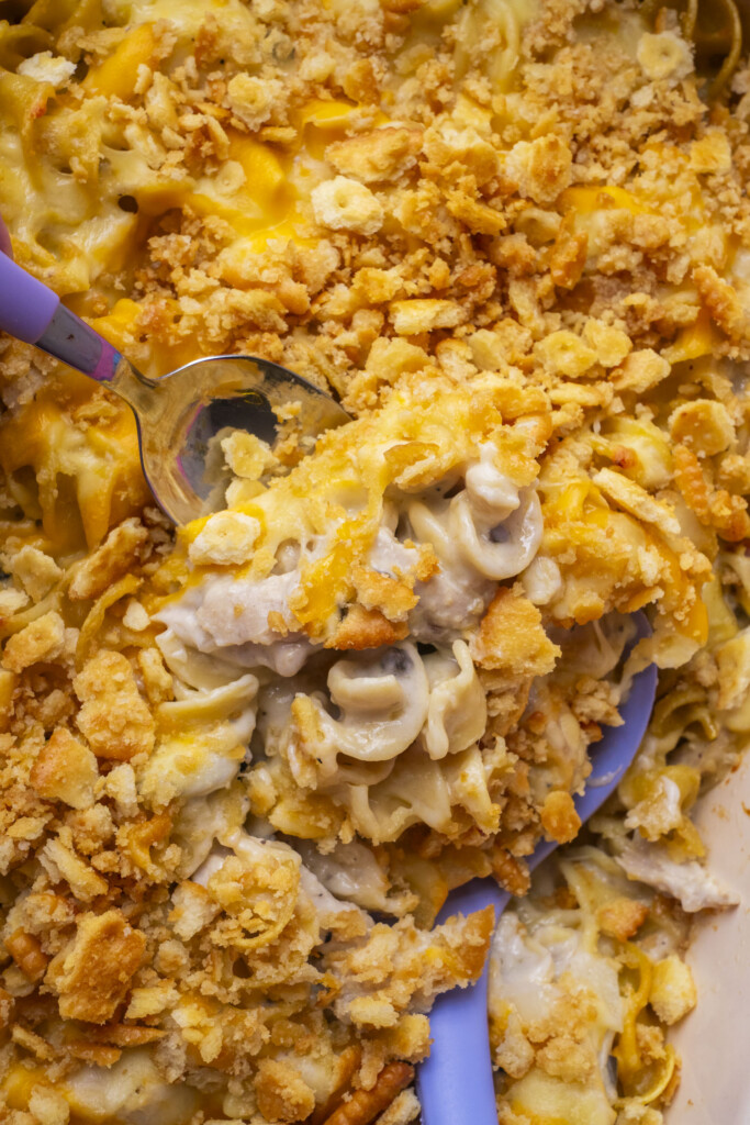 A close-up of Million Dollar Chicken Casserole with noodles, chunks of chicken, and a golden, crumbly cracker topping. Two serving spoons are scooping out a portion from the dish.