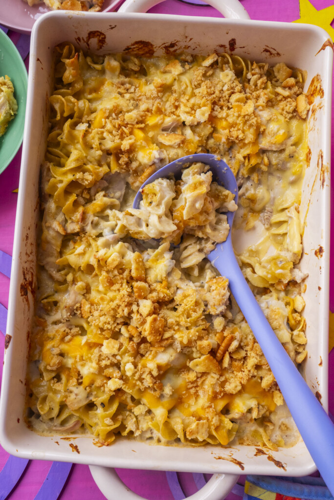 A baking dish filled with creamy Million Dollar Chicken Casserole topped with golden, crushed crackers and melted cheese. A blue serving spoon is scooping out a portion. The dish is set on a pink surface.