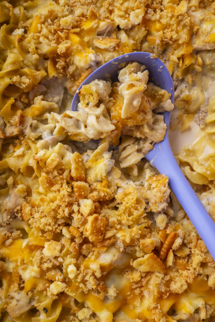 Close-up of a cheesy Million Dollar Chicken Casserole topped with golden cracker crumbs, with a blue serving spoon scooping out a portion, revealing creamy pasta underneath.