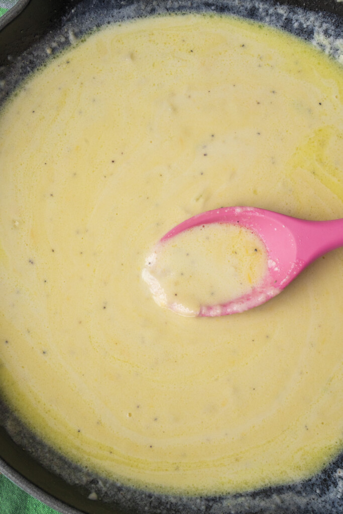A pink spoon stirs a creamy yellow sauce with visible black pepper specks in a pan.