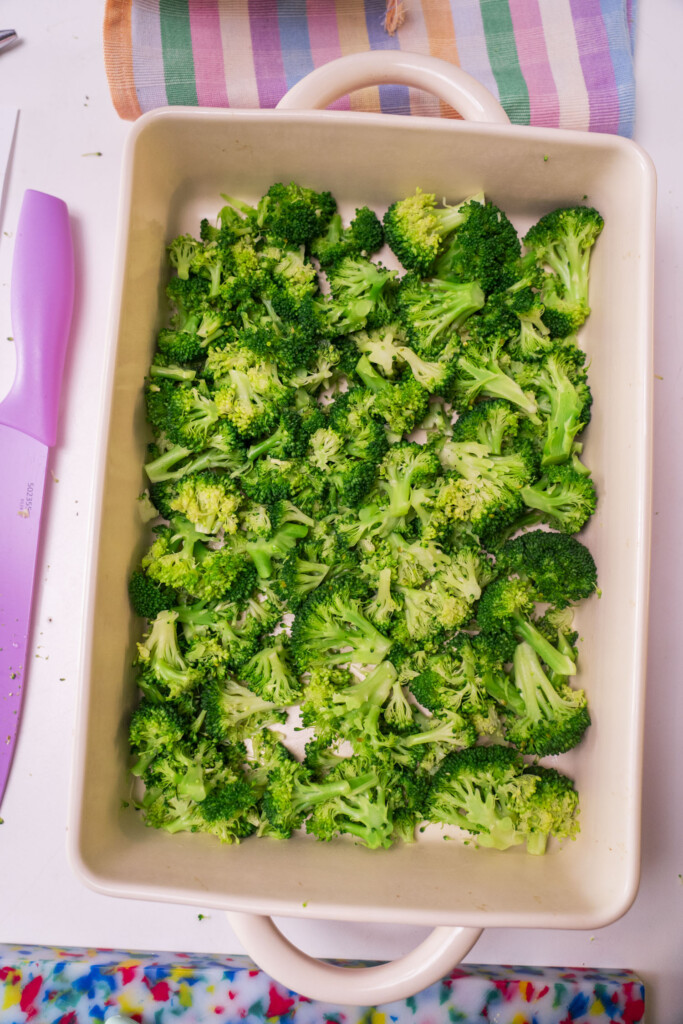 A baking dish filled with fresh broccoli florets sits on a white surface. Nearby are a pink knife, a striped towel, and a colorful mat, adding vibrant touches to the cooking scene.