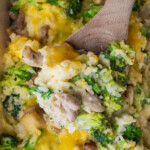 A close-up of a creamy casserole with broccoli, chicken pieces, rice, and melted cheese being scooped with a wooden spoon.