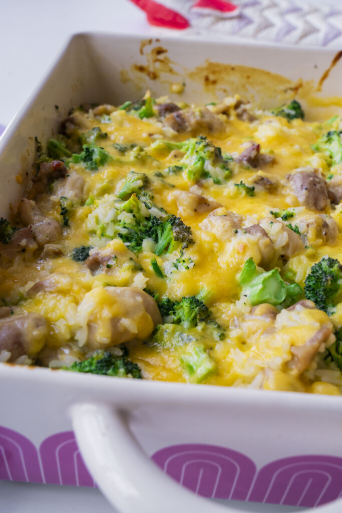 A close-up of a baked casserole in a white dish, featuring melted cheese, broccoli, rice, and pieces of chicken. The dish is colorful and savory, with a golden, bubbly cheese topping.