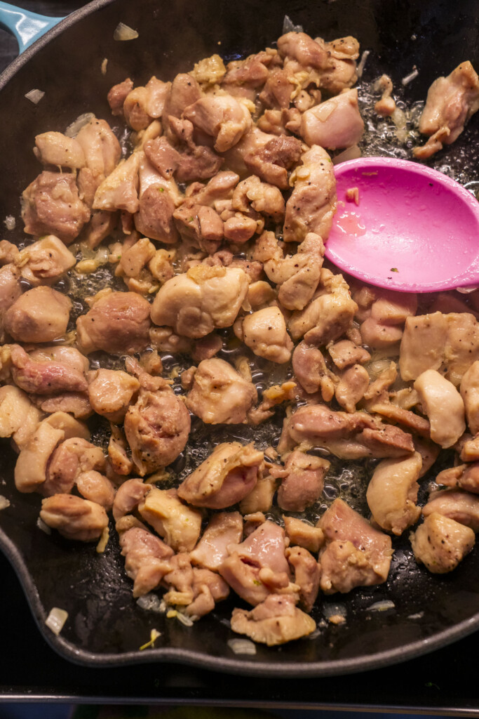 Chopped pieces of chicken thighs cooking in a black skillet with diced onions, next to a pink spatula.